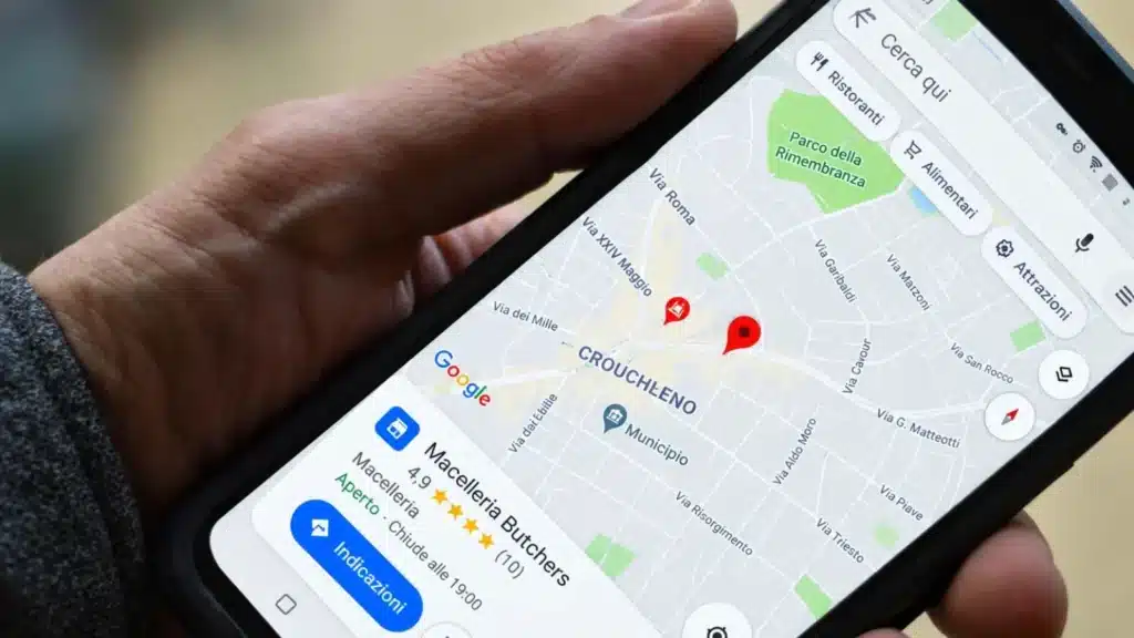Smartphone tenuto in mano che mostra l'applicazione di mappe con i risultati di ricerca geolocalizzata e la scheda di un'attività commerciale locale.